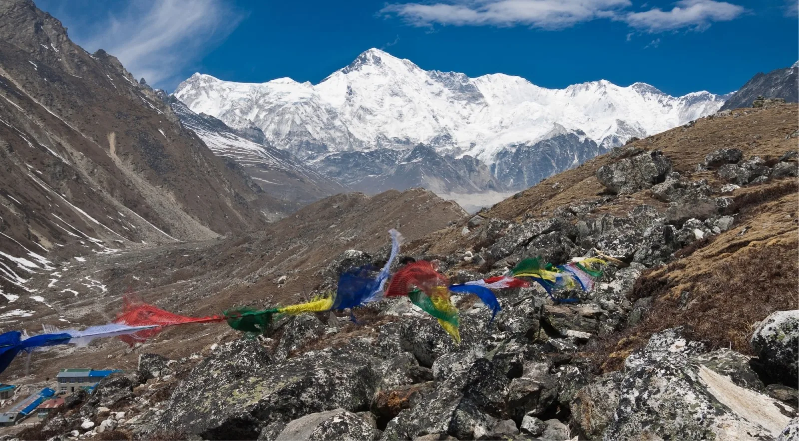 Prayer flags snapping in the wind above a boulder-strewn ridge with a snow massif and lodges beyond