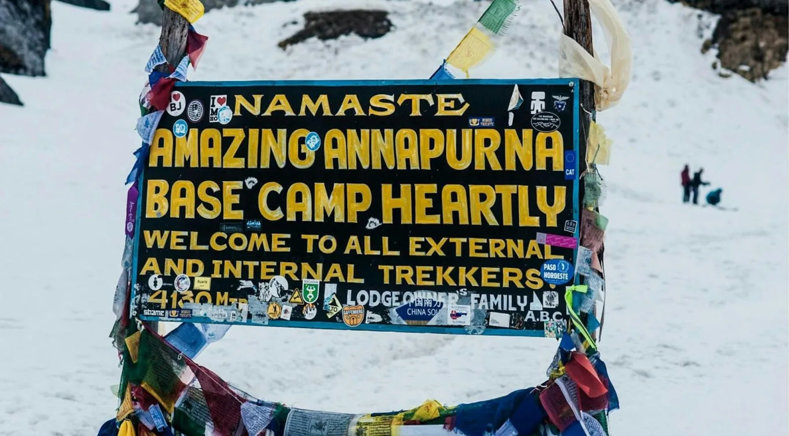 Sticker-covered welcome board reading Namaste Amazing Annapurna Base Camp 4130 Mtr, framed by prayer flags