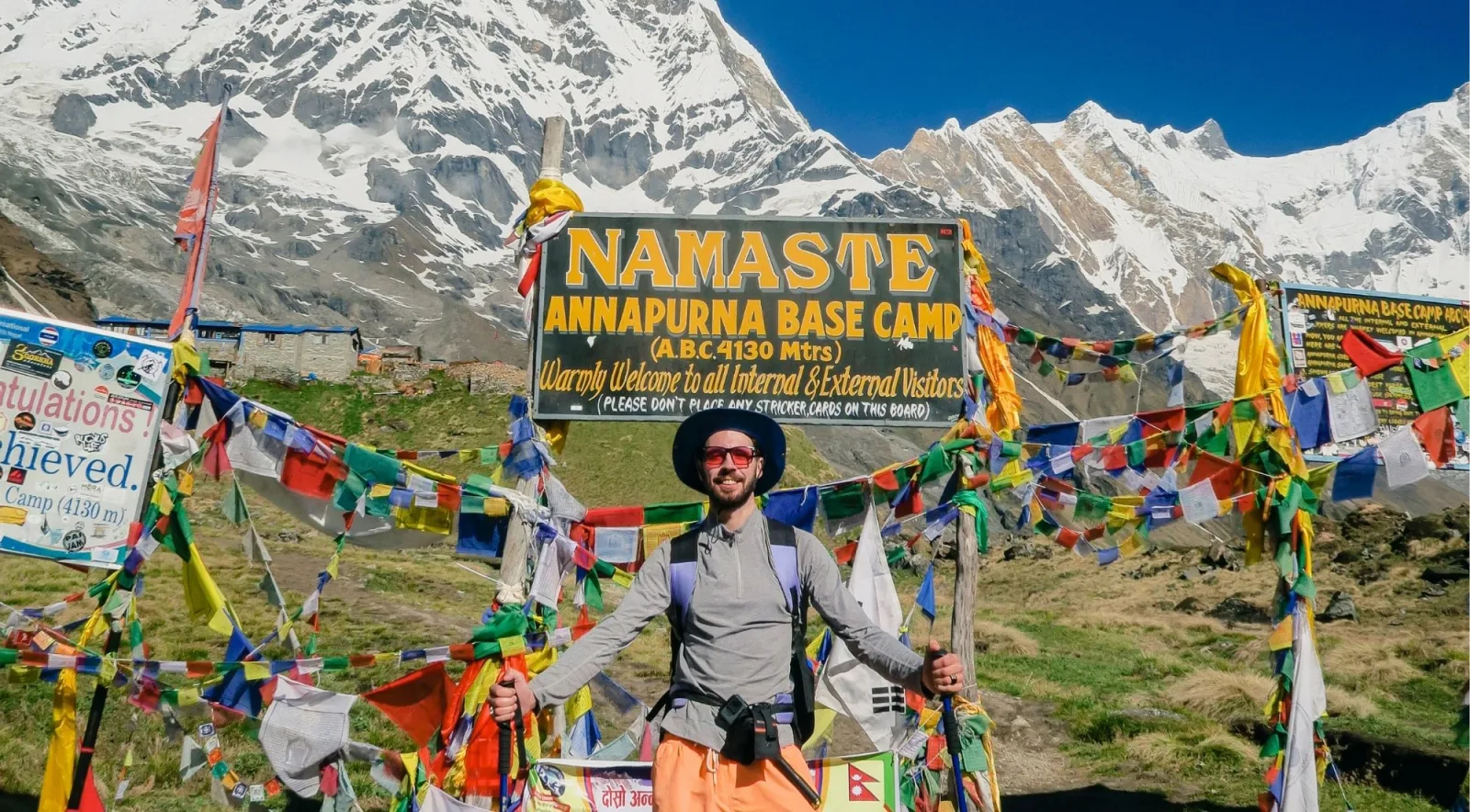 Smiling trekker with poles standing among prayer flags at the Annapurna Base Camp sign on green grass