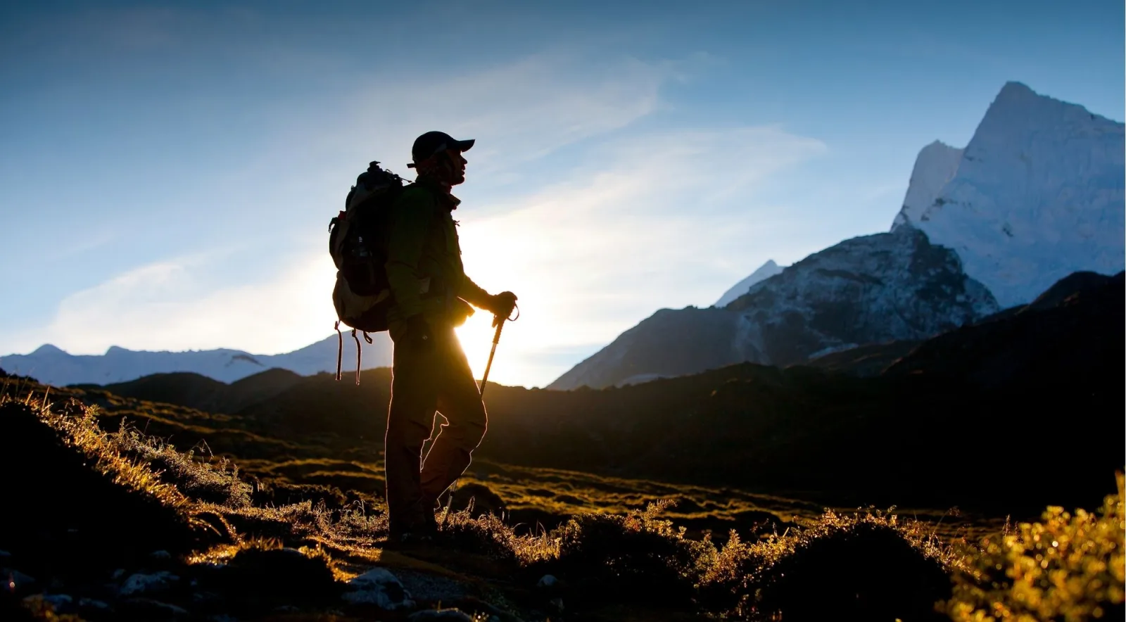 Trekker silhouetted at golden hour with a backpack and poles, a sharp Himalayan peak on the skyline