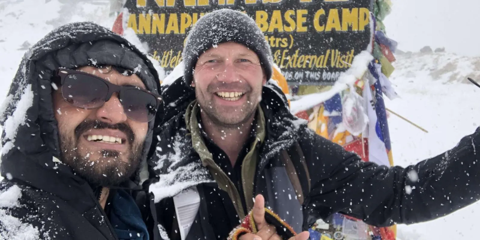 Two trekkers grinning in falling snow in front of the Annapurna Base Camp sign